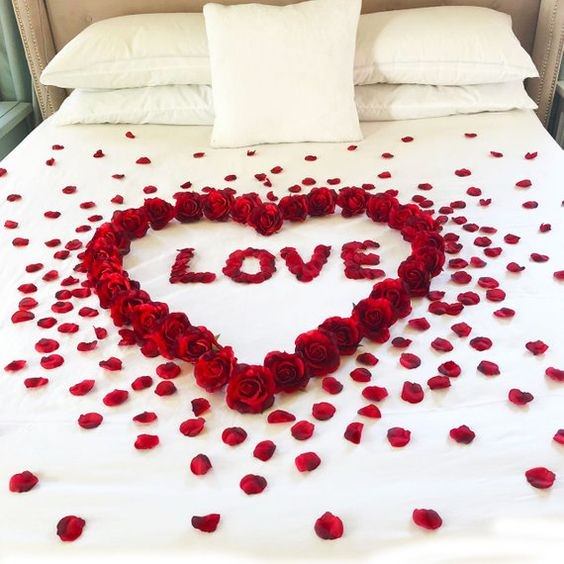 تخت خواب عروس و دامادی که روی روتختی سفید آن گل و گلبرگ های رز قرمز به شکل قلب و love قرار داده اند